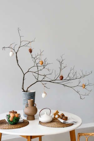 Basket with Easter eggs, teapot and vase with tree branches on dining table near light wall