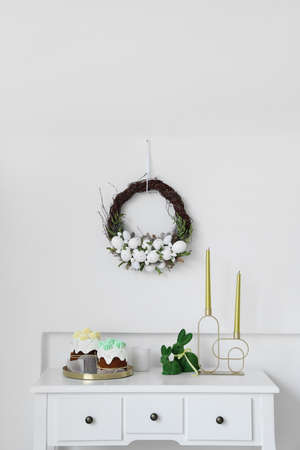Table with Easter cakes, rabbits and candles near light wall with wreath