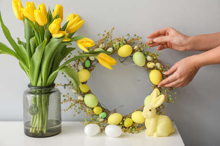 Female hands with Easter Wreath, eggs, rabbit and tulips near gray wall