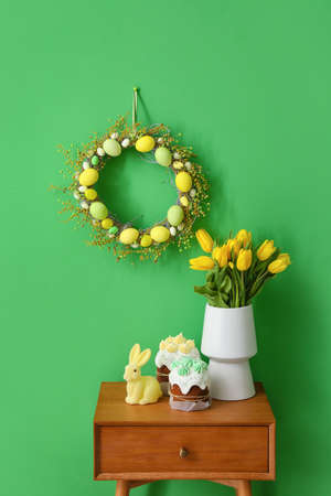 Easter cakes, rabbit and vase with tulips on table near color wall with wreath
