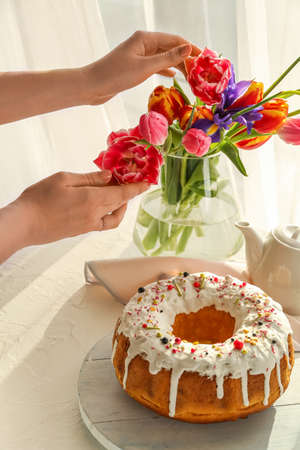 Woman with flowers and tasty Easter cake on table in room