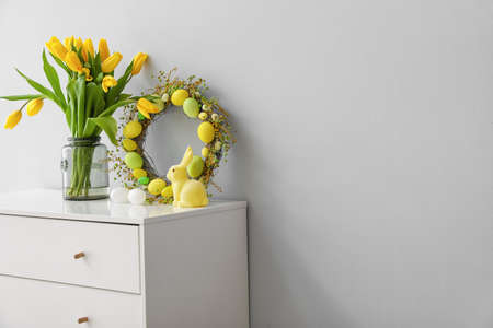 Easter wreath, rabbit and vase with tulips on chest of drawers near gray wall