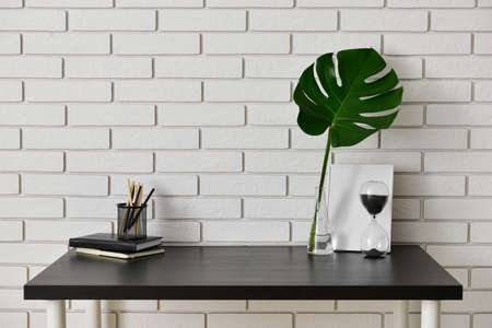 Stylish workplace with hourglass, stationery and vase with tropical leaf near light brick wall