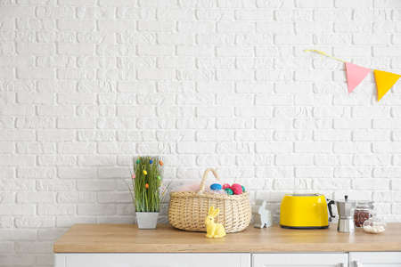 Basket with Easter eggs, stylish decor and different utensils on kitchen counter near white brick...