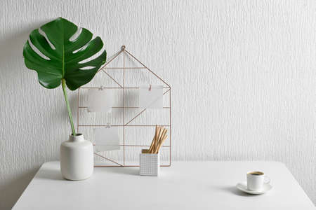 Workplace with palm leaf in vase, sticky notes and cup of coffee near light wall