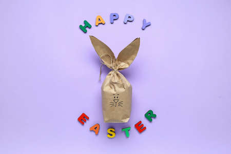 Text HAPPY EASTER made of colorful letters and bunny gift bag on lilac background