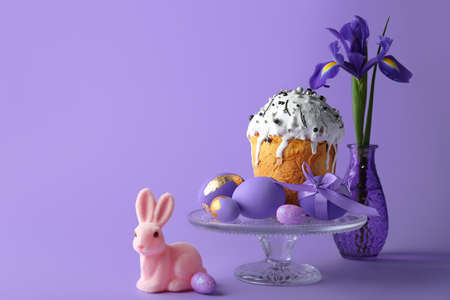 Cute Easter bunny, cake, painted eggs and vase with flowers on purple backgroundの写真素材