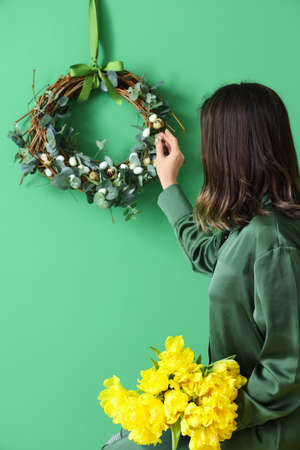 Young woman with bouquet of tulips touching Easter wreath on color wallの写真素材