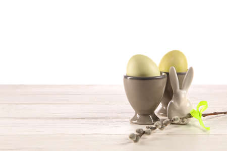 Cups with painted Easter eggs, pussy willow and figurine of bunny on table against white background