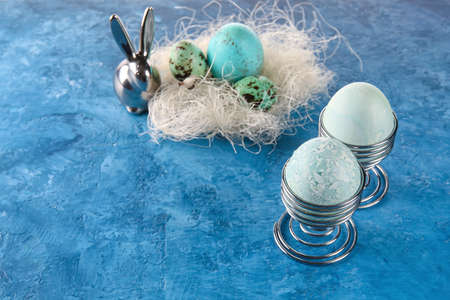 Cups with painted Easter eggs and figurine of bunny on blue background