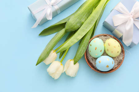 Bowl with painted Easter eggs, tulip flowers and gift boxes on blue background