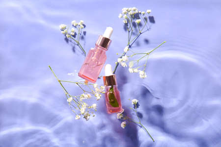 Bottles with natural serum and gypsophila flowers in water on lilac background