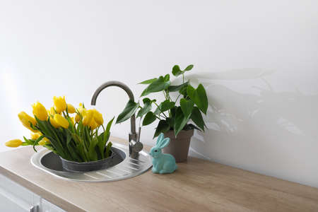Bucket with fresh tulip flowers in sink, houseplant and figurine of bunny on table in kitchen....