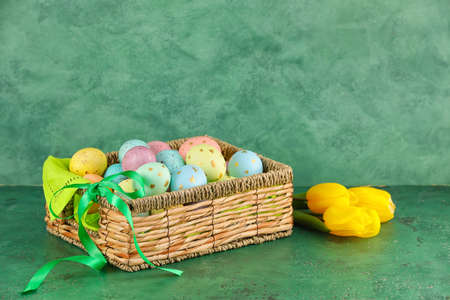 Wicker basket of painted Easter eggs and flowers on green background