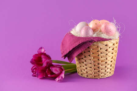 Wicker basket of painted Easter eggs and flowers on color background