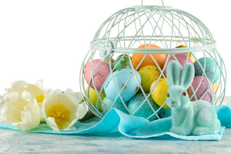 Basket of painted Easter eggs, toy bunny and flowers on table against white background