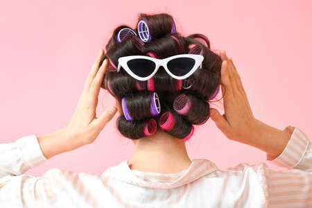 Beautiful young woman in pajamas, hair curlers and sunglasses on pink background, back viewの写真素材