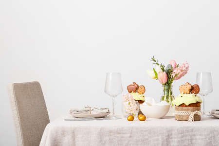Beautiful table setting with Easter cakes, eggs and flowers near light wall