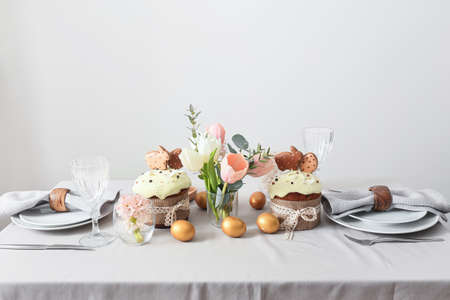 Beautiful table setting with Easter cakes, eggs and flowers near light wall