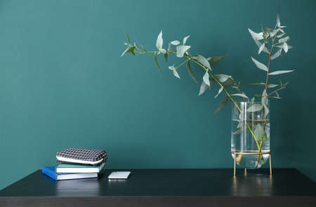 Workplace with plant branches in vase, pencil case and books near green wall