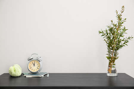 Workplace with plant branches in vase, alarm clock and notebooks near light wall