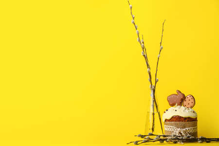 Vase with willow branches and Easter cake decorated with cookies on yellow background
