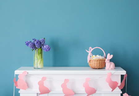 Vase with beautiful flowers, Easter eggs in basket and bunny on mantelpiece near color wallの写真素材