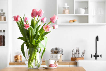 Vase with tulips, cup and macarons on table in light kitchenの写真素材
