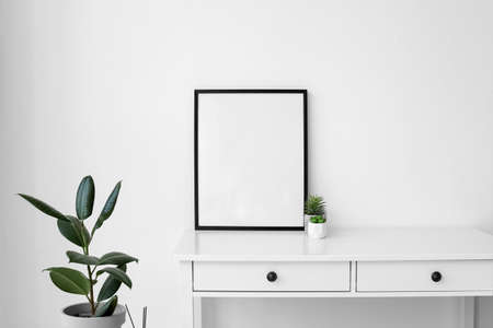 Big blank frame and flowerpots on table near light wall