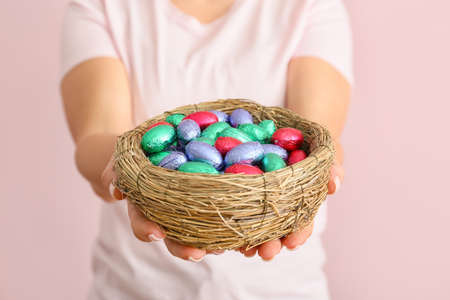 Woman holding nest with chocolate eggs on color background, closeup
