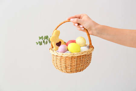 Woman holding gift basket with painted Easter eggs and cookies on light background