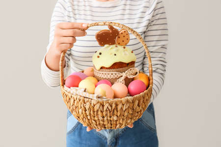 Woman holding gift basket with painted Easter eggs and cake on light background, closeup