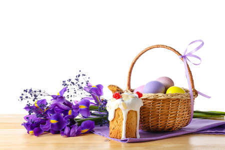 Gift basket with painted Easter eggs, flowers and cake on table against white background