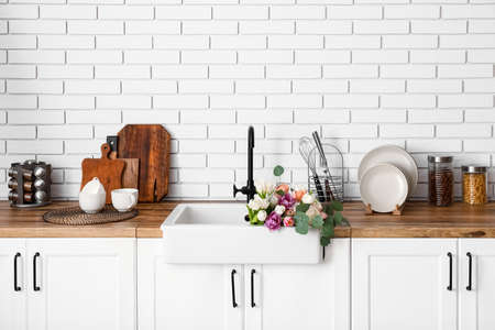 Sink with beautiful flowers and kitchen utensils on counters near white brick wallの写真素材