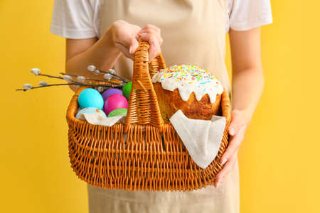 Woman holding gift basket with painted Easter eggs, willow branches and cake on yellow background