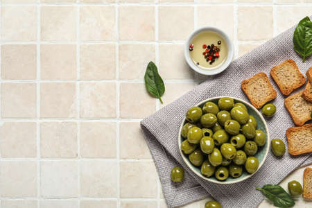 Composition with bowl of tasty green olives, oil and crackers on light tile