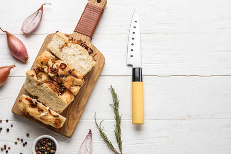 Board with tasty Italian focaccia, onion, spices and knife on light wooden background