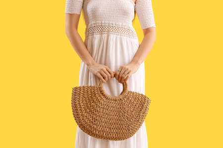 Elegant woman with stylish jewelry holding rattan handbag on yellow backgroundの写真素材