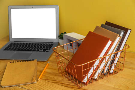 Metal basket with books, clock and laptop on table near yellow wallの写真素材