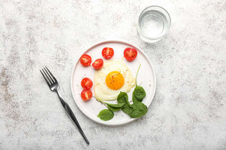 Plate with tasty fried egg, tomatoes, spinach and glass of water on light background