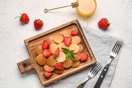 Wooden board with mini pancakes, delicious strawberry and honey on light background