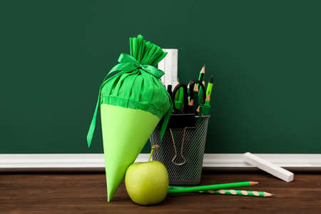 Holder with stationery, school cone and apple on table near chalkboard