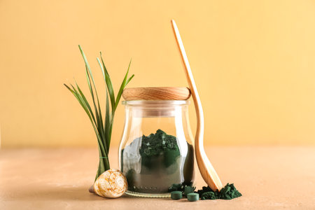 Glass jar of spirulina powder, spoon, pills, seashell and leaves on table against yellow backgroundの写真素材