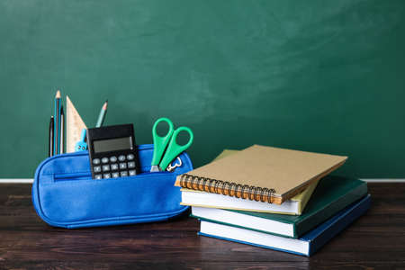 Pencil case with school stationery and books on table near green chalkboard