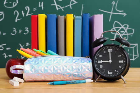 Pencil case with school stationery, alarm clock and apple on table near green chalkboard