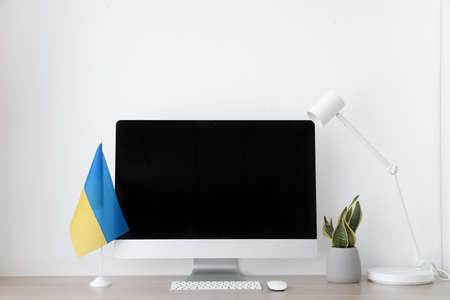 Modern workplace with computer, flag of Ukraine, houseplant and lamp near light wallの写真素材