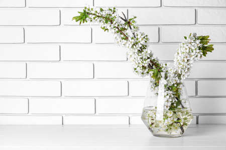 Vase with beautiful blooming tree branches on wooden table near white brick wallの写真素材