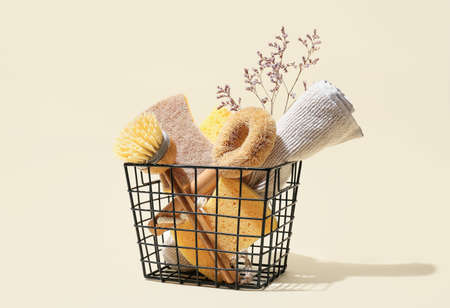 Basket with set of cleaning supplies, towel and flowers on color background
