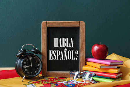 Chalkboard with text HABLA ESPANOL?(DO YOU SPEAK SPANISH?), flag, clock and stationery on table against color backgroundの写真素材