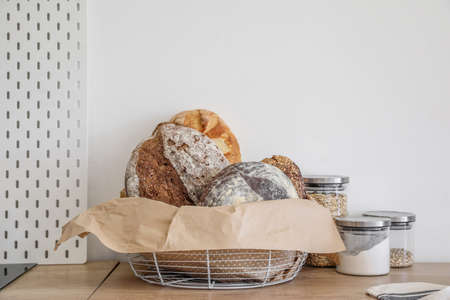Basket with loaves of fresh bread, parchment paper and jars on kitchen counter near light wallの写真素材